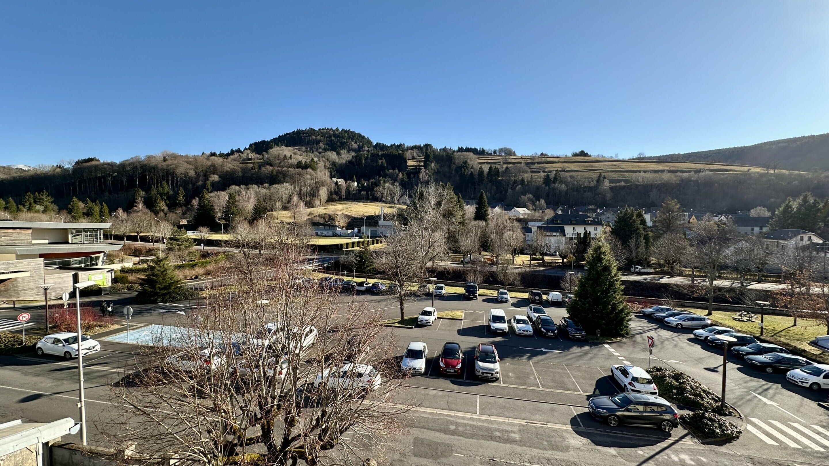 A parking lot with multiple cars, a building with a modern design, and a hilly landscape in the background.