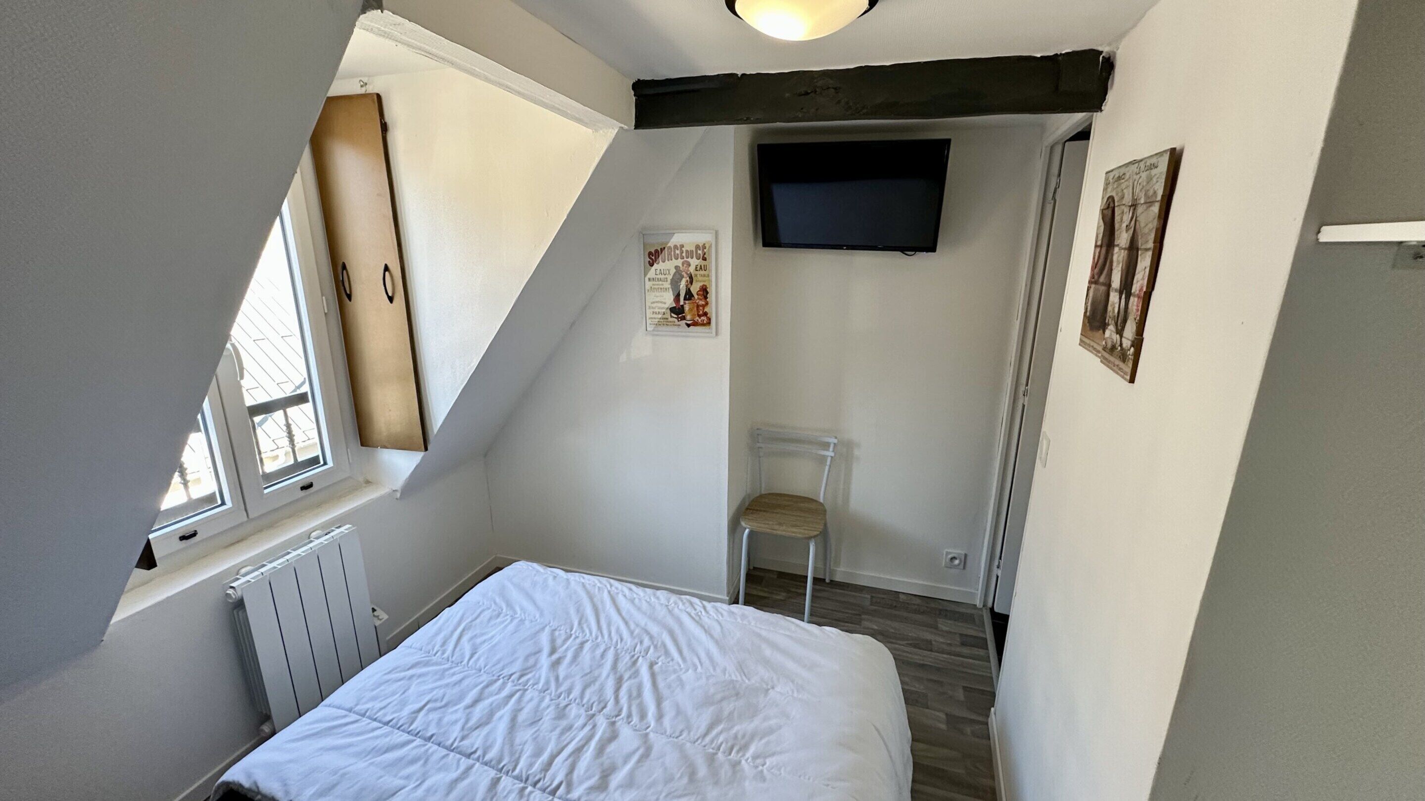 Een kleine, goed verlichte kamer met een schuine kap, een bed, een stoel, een radiator en een aan de muur bevestigde televisie.