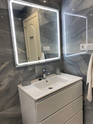 A bathroom with a white vanity, a mirror with LED lighting, and a marble wall. - Marbella Real Apartment (Marbella)