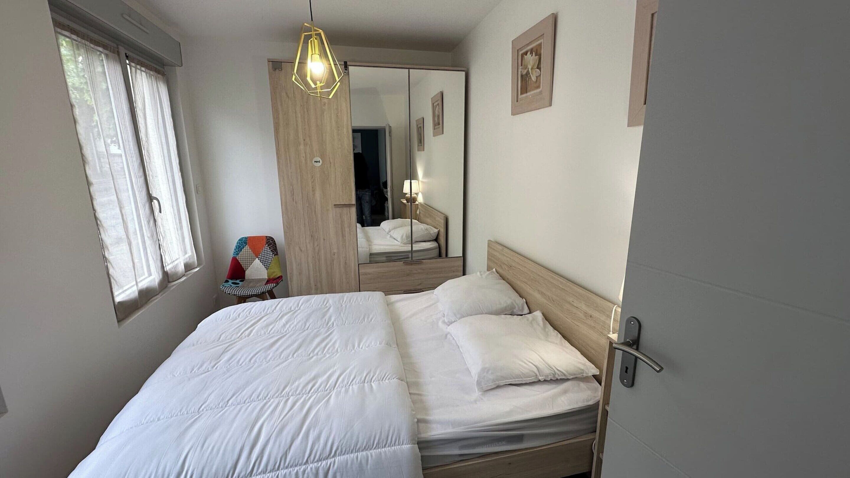 A neatly made bed with white linens, a wooden headboard, and a mirror reflecting a room with a chair and a lamp.