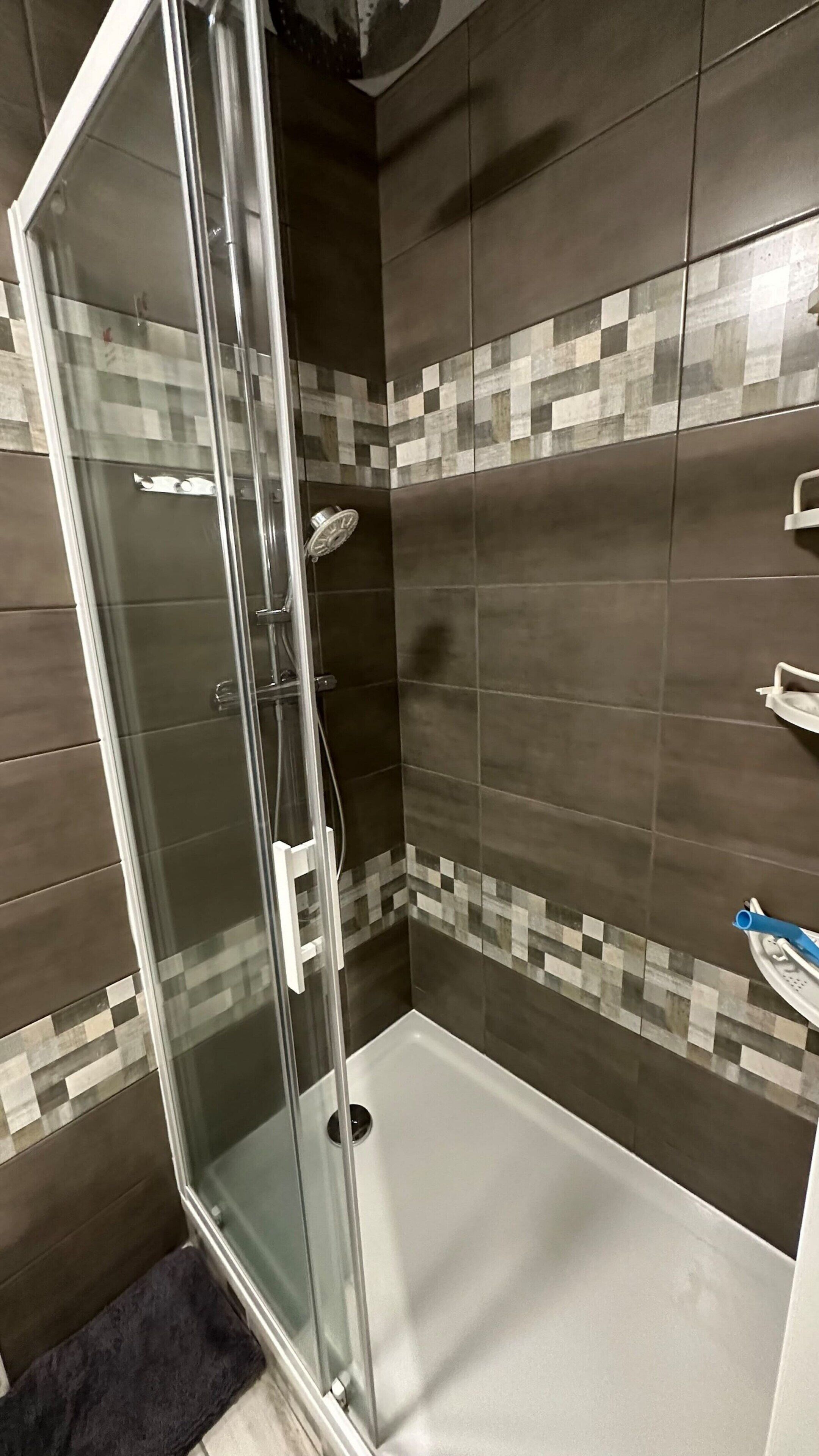 A glass-enclosed shower with a tiled wall, a white floor, and a dark brown tiled shower area.