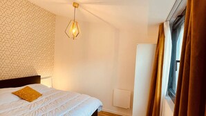 Quarto com cama, parede com padrão geométrico, luminária pendente, janela com cortinas e radiador.
