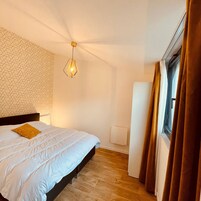 Un dormitorio con una cama, un panel de pared con estampado geométrico, una lámpara colgante, una ventana con cortinas y un radiador.