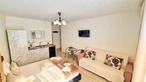 A modern, well-lit room with a bed, a sofa, a dining table, and a small kitchen area.