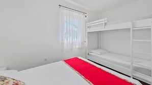 Een kamer met een stapelbed, een enkel bed met een rode loper en een raam met dunne gordijnen.