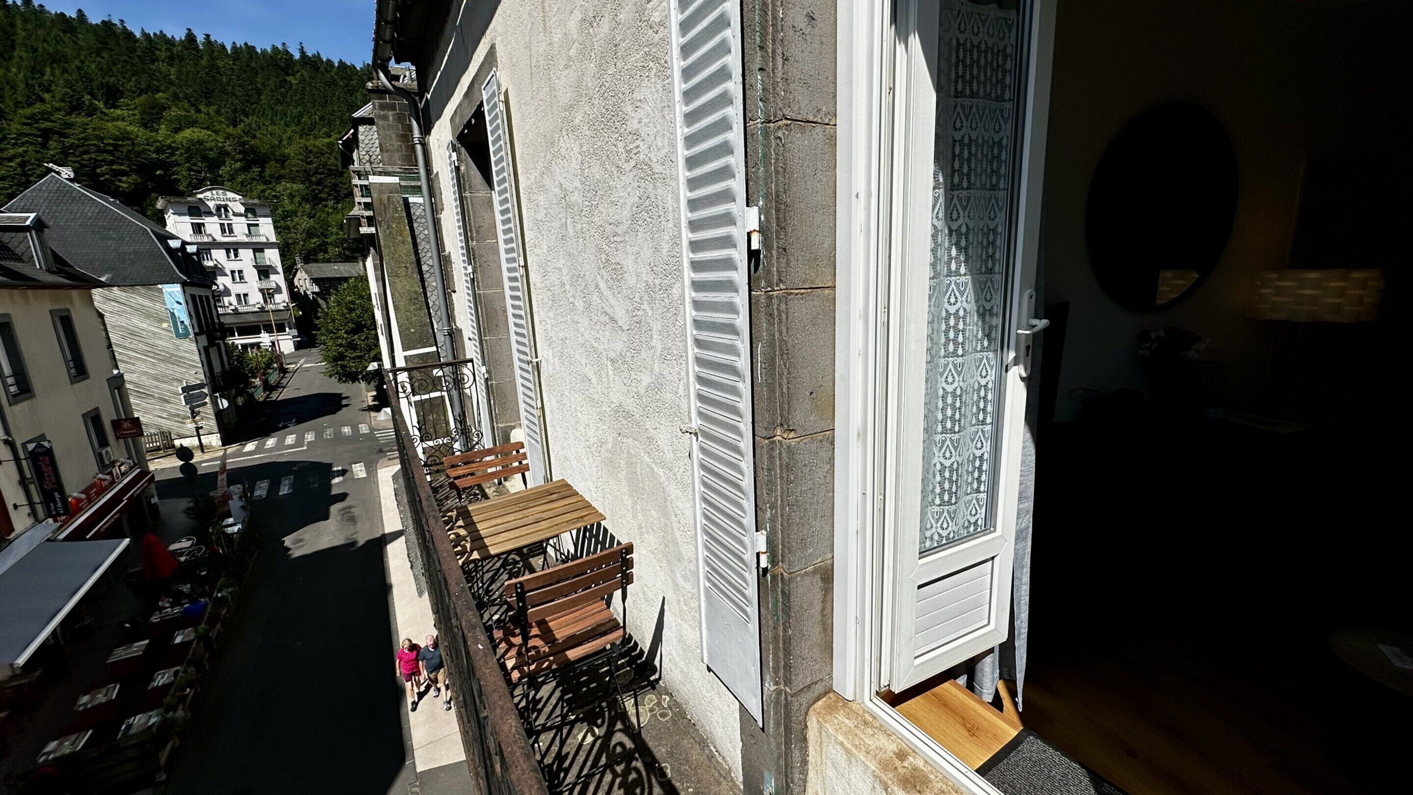 En smal balkong med en träbänk, med utsikt över en gata med byggnader och en skogbeklädd kulle i bakgrunden.