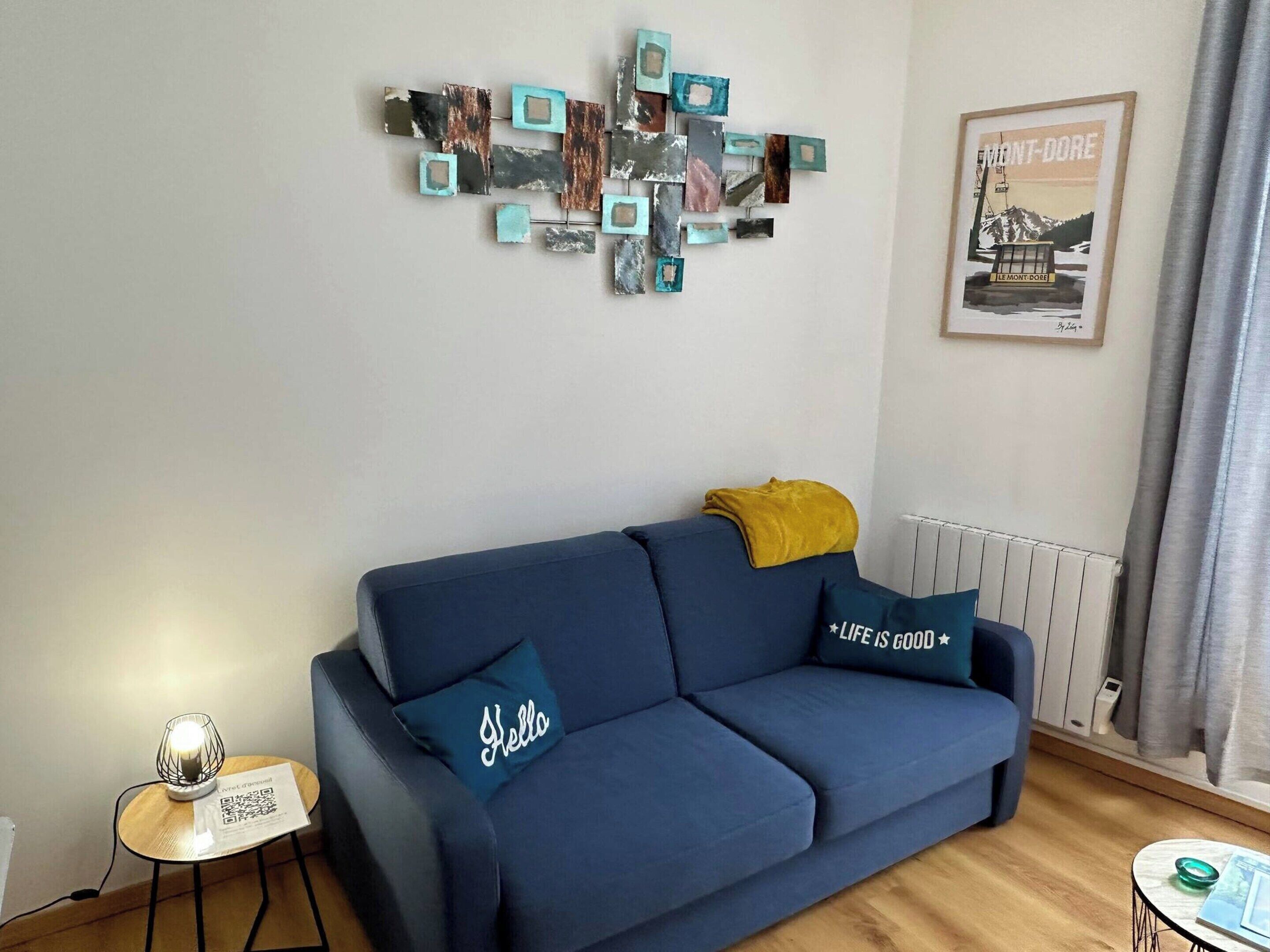 A living room with a blue sofa, a decorative wall piece, a framed picture, and a small table with a lamp and a QR code.