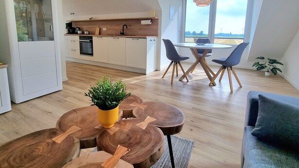 Eine moderne Küche mit weißen Schränken, ein Essbereich mit rundem Tisch und Stühlen sowie ein Blick ins Freie durch große Fenster.