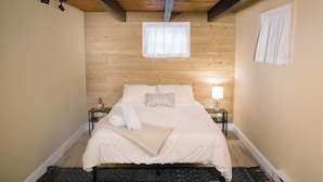 A bedroom with a wooden ceiling, a bed with a white bedspread, a nightstand with a lamp, and a window with a white curtain.