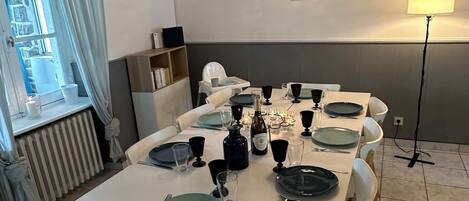 A dining area with a white table set for six, surrounded by wooden chairs. A radiator is visible to the left, and a window with curtains is on the left side.