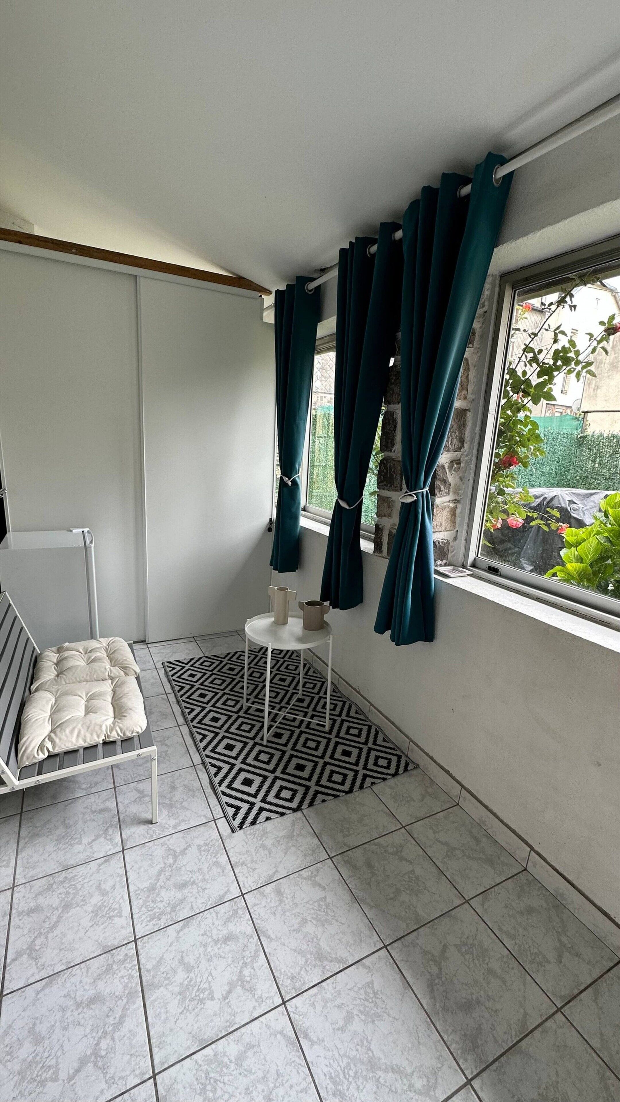 A room with a bed, a small table with two pitchers, a patterned rug, and a window with curtains.