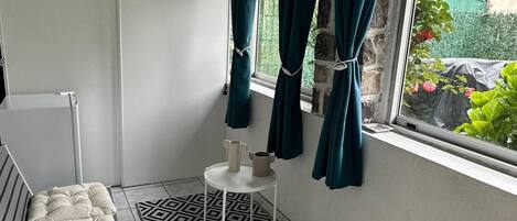 A room with a bed, a small table with two pitchers, a patterned rug, and a window with curtains.