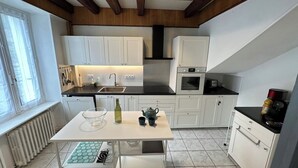 A compact kitchen with white cabinets, a black countertop, a built-in oven, and a window with blinds. - Centrally Located Villa in Mont-dore (La Bourboule)