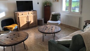 A living room with a television, wooden cabinet, two round tables, a sofa, and a window with a view of greenery. - Large, spacious Germaine house in a quiet location 10 minutes from the sea. (Sibiril)