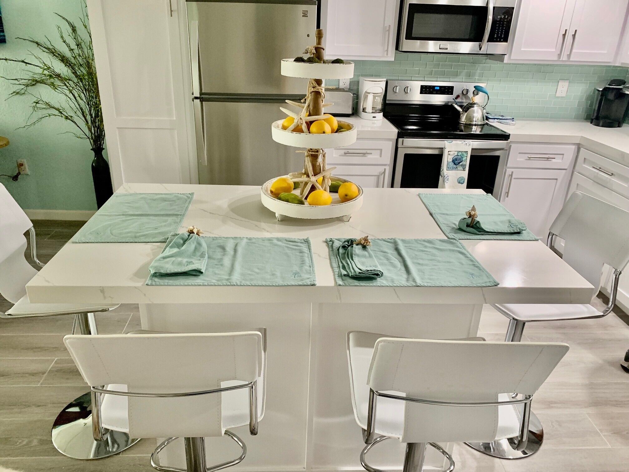 A modern kitchen with a stainless steel refrigerator, a microwave, and a stove. There is a fruit bowl on the counter and a potted plant in the corner.