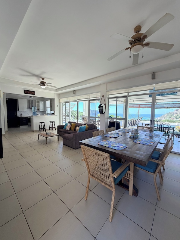 A spacious living area with a dining table, wicker chairs, and a sofa. There is a ceiling fan and a view of the ocean through large windows.