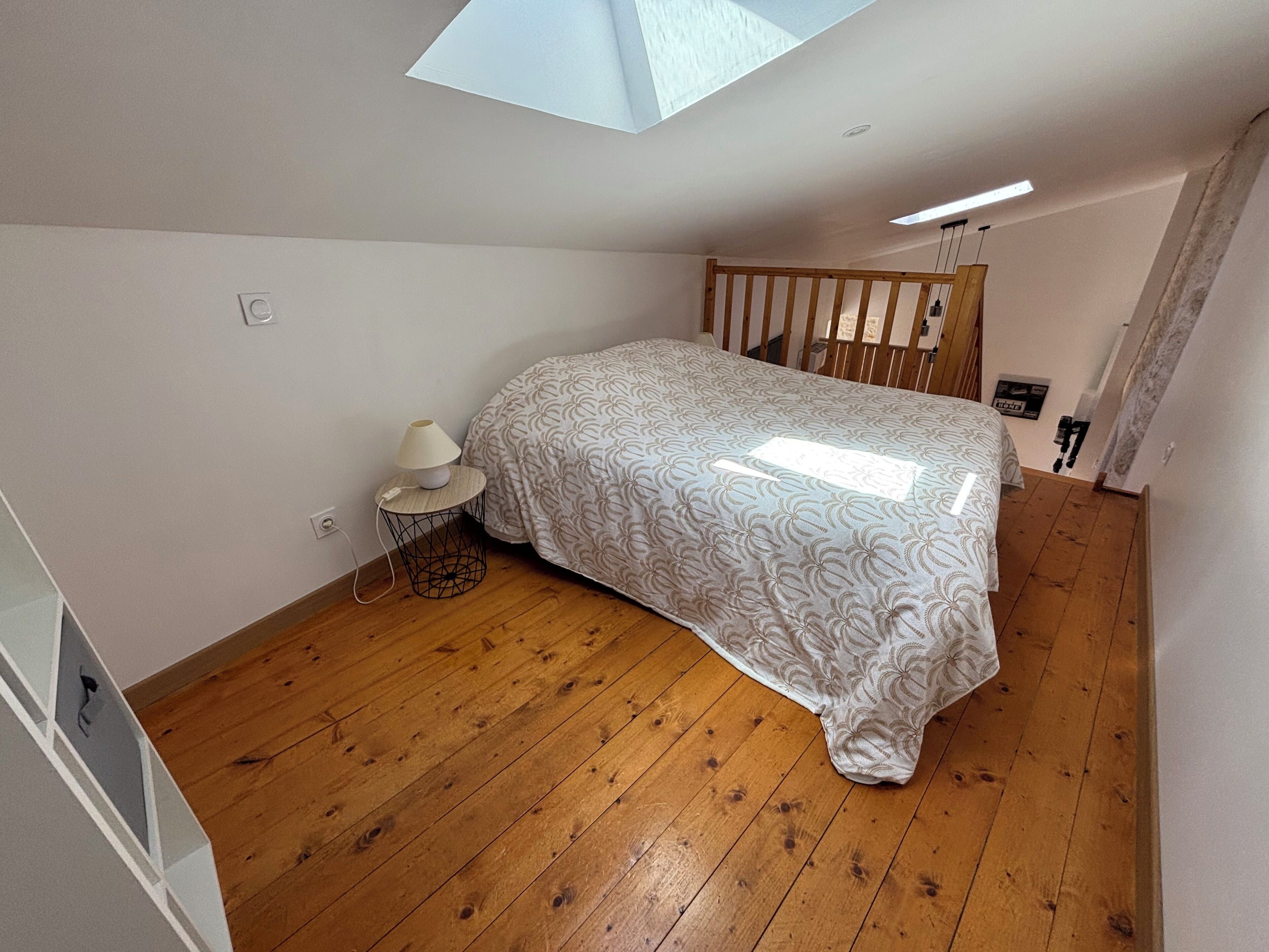 A bedroom with a bed, a bedside table with a lamp, a wooden floor, and a skylight.