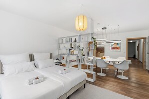 Ein modernes Hotelzimmer mit einem großen Bett, weißen Möbeln, einem Essbereich mit Tisch und Stühlen sowie einem Bücherregal mit verschiedenen Gegenständen.