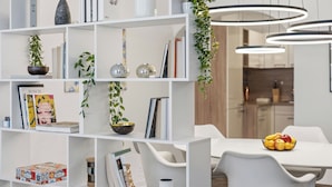 Un interior moderno con muebles blancos, una estantería con libros y objetos decorativos, y un comedor con frutero.