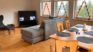 A modern living room with a grey sectional sofa, a flat-screen TV, a dining table with placemats, and windows with triangular wooden frames. - Haus De Kleene & Gruusse in Mildenau (Mildenau)