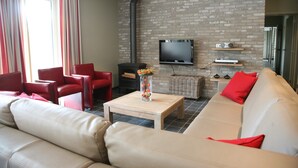 A living room with a sofa, red chairs, a TV mounted on a brick wall, and a fireplace. - Countryside Holiday Home (Alveringem)