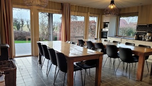 A spacious dining area with a large table and chairs, large windows with curtains, and a kitchen area with modern appliances. - Countryside Holiday Home (Alveringem)