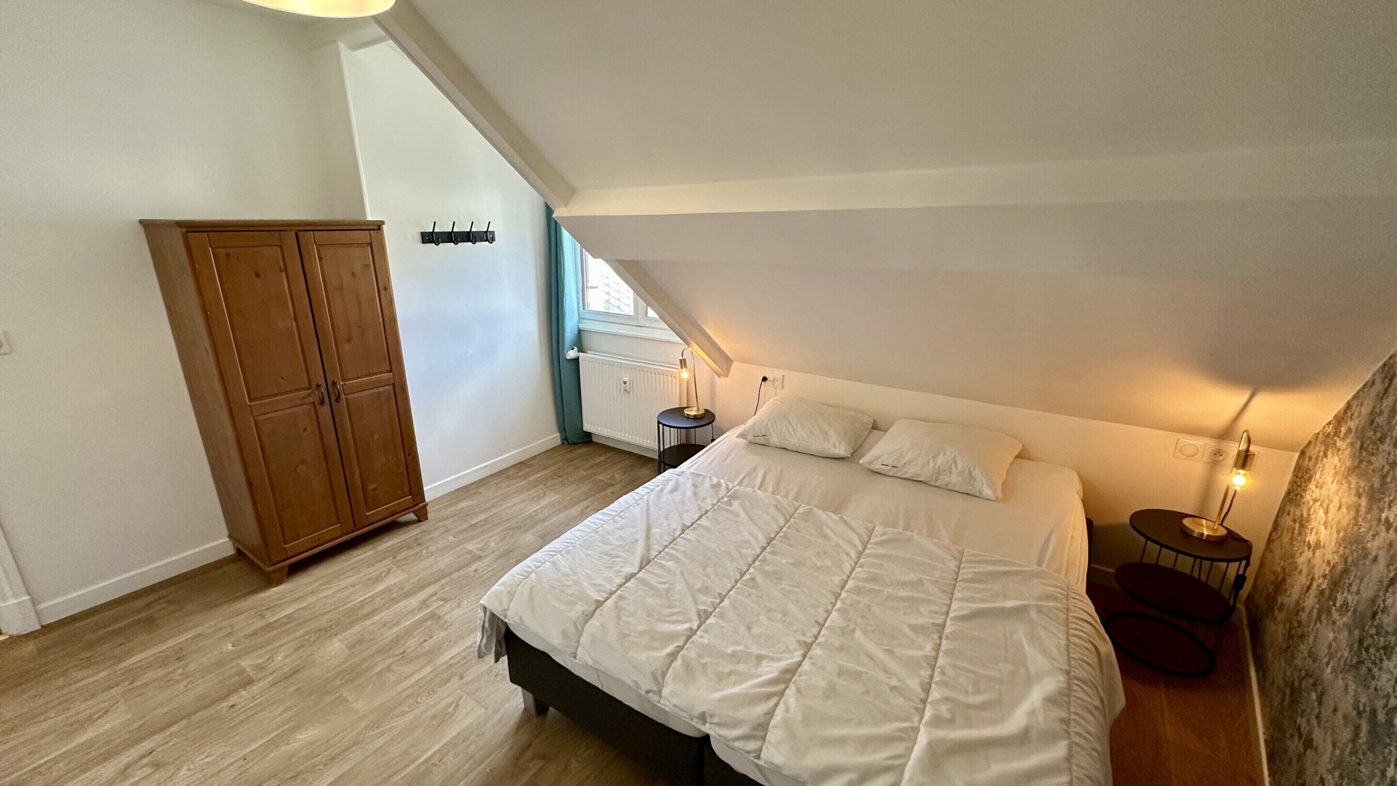 Une chambre à coucher avec un grand lit, une armoire, une petite table de chevet avec une lampe et une fenêtre avec des rideaux.