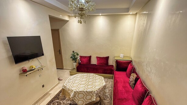 A modern living room with a flat-screen TV, a red sofa, a patterned rug, and a chandelier. - 2 balcony rooms in Mabrouka  5 minutes from Guéliz (Marrakech)