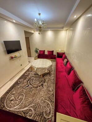 A modern living room with a flat-screen TV, a red sofa, a patterned rug, and a chandelier. - 2 balcony rooms in Mabrouka  5 minutes from Guéliz (Marrakech)