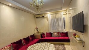A living room with a sofa set, a round coffee table, a chandelier, a flat-screen TV, and a patterned rug. - 2 balcony rooms in Mabrouka  5 minutes from Guéliz (Marrakech)