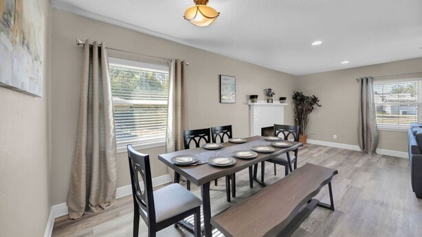 Un coin repas avec une longue table, des chaises et un banc. Les fenêtres sont dotées de rideaux, et on y trouve un luminaire au plafond ainsi qu’une cheminée.