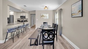 Un espace repas avec une longue table, des tabourets de bar et une cuisine en arrière-plan.
