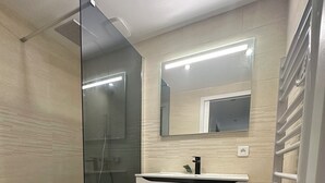 Un baño moderno con cabina de ducha de vidrio, tocador blanco con espejo y un pequeño cubo de basura.