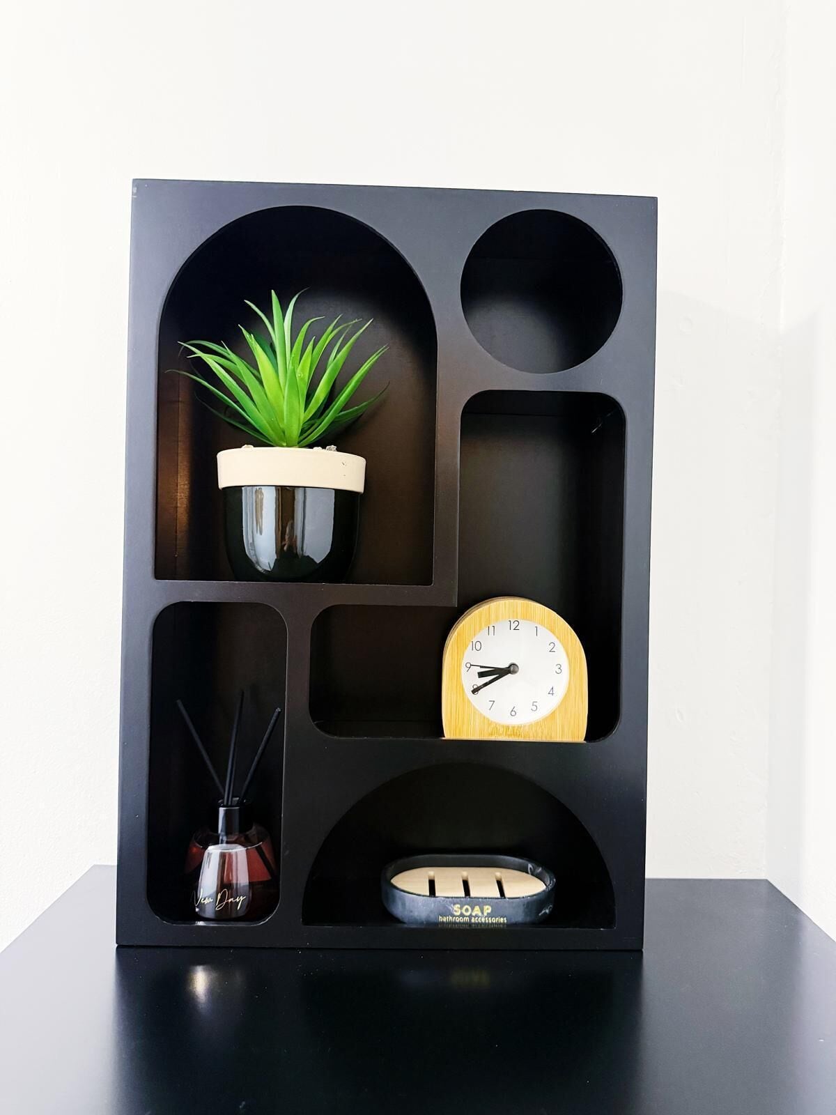 A black shelf with multiple compartments containing a potted plant, a clock, a small vase, and a soap dish.