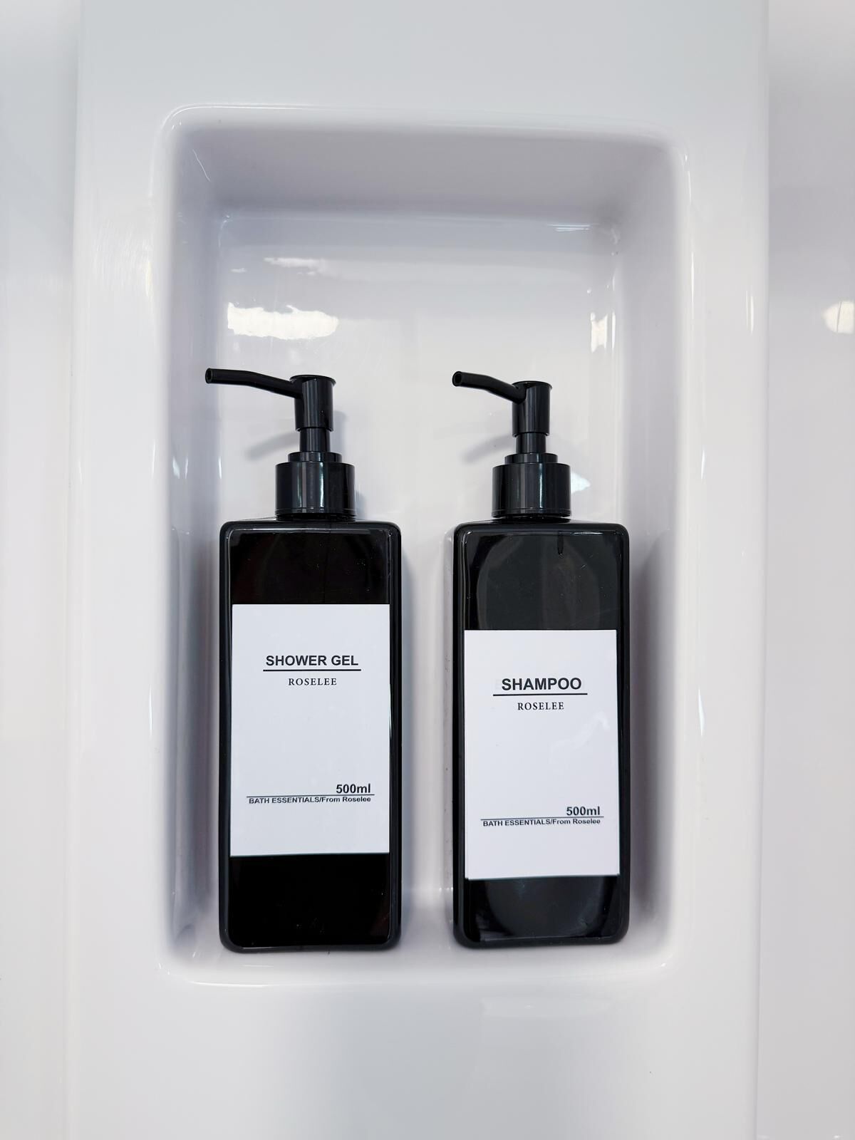 Dos botellas negras de productos de baño Roselee, una etiquetada como 'Gel de ducha' y la otra 'Champú', cada una con dispensador de bomba y una etiqueta que indica un volumen de 500 ml.