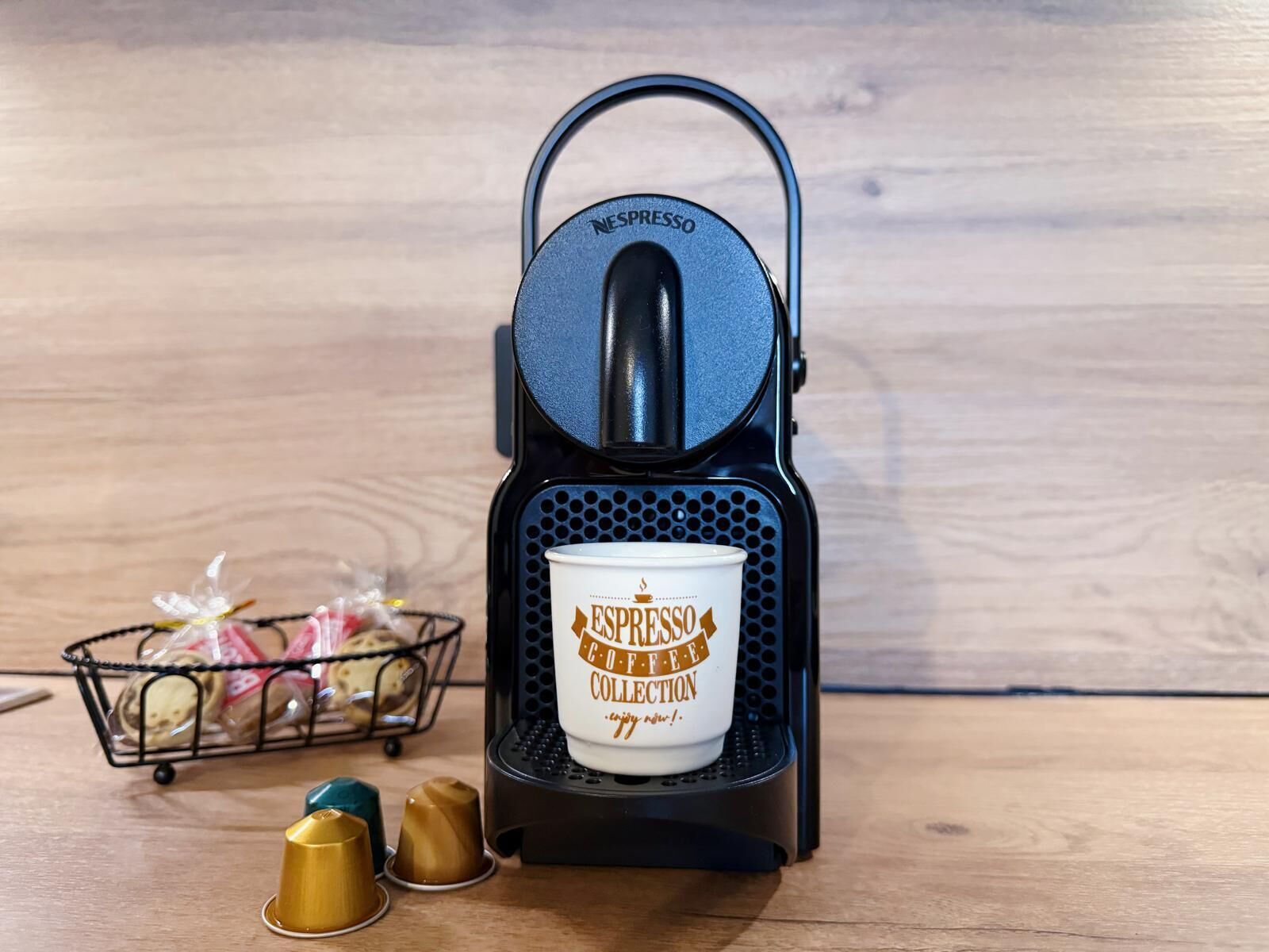 Une machine à café Nespresso, accompagnée d’une tasse et de trois gobelets à café, le tout posé sur une surface en bois.
