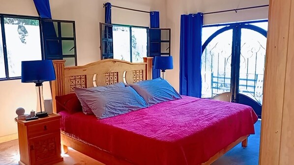 Ein Bett mit Holz-Kopfteil, einer roten Tagesdecke und zwei Kissen. Ein Nachttisch mit Lampe und ein kleiner Tisch ebenfalls mit einer Lampe. Das Zimmer verfügt über ein Fenster mit blauen Vorhängen und Blick ins Freie.