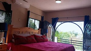 Une chambre à coucher avec un grand lit, une tête de lit en bois et une vue par une fenêtre ornée d’une grille en métal.