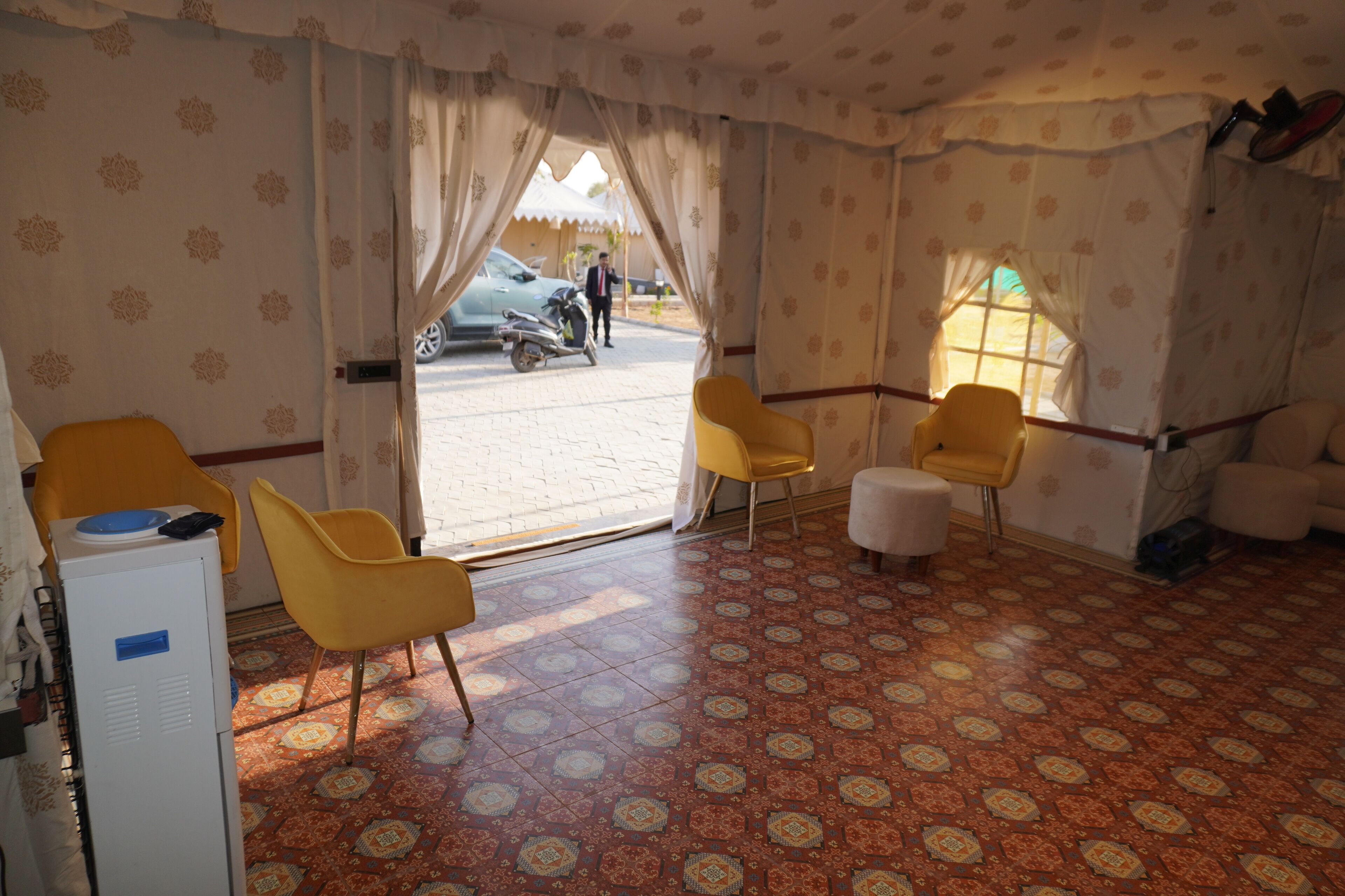 A room with patterned carpet, yellow chairs, a white refrigerator, and a view of a street with a motorcycle and a car.