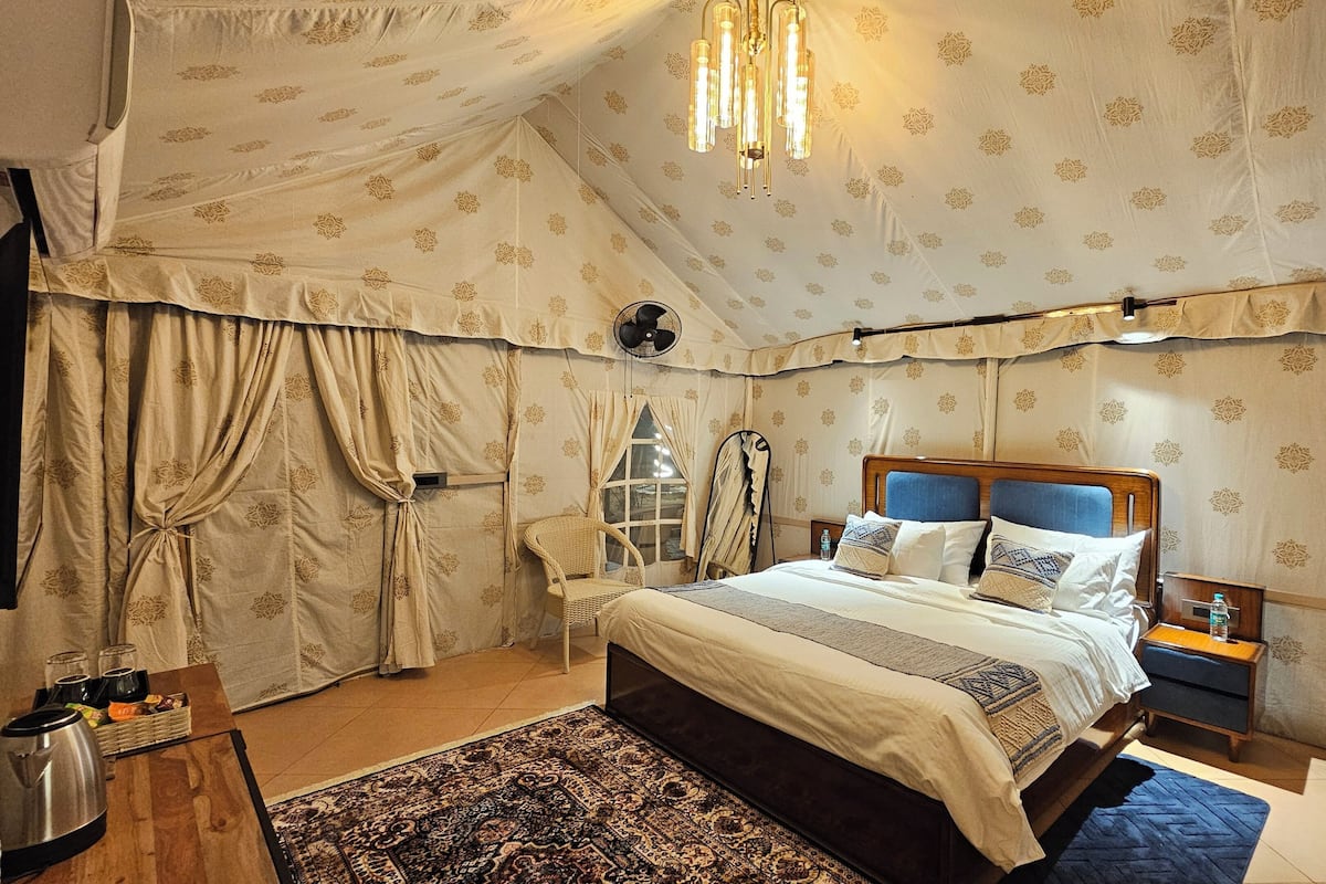 Una habitación con carpa, una cama, una mesita de noche, una lámpara, una silla y una alfombra estampada.