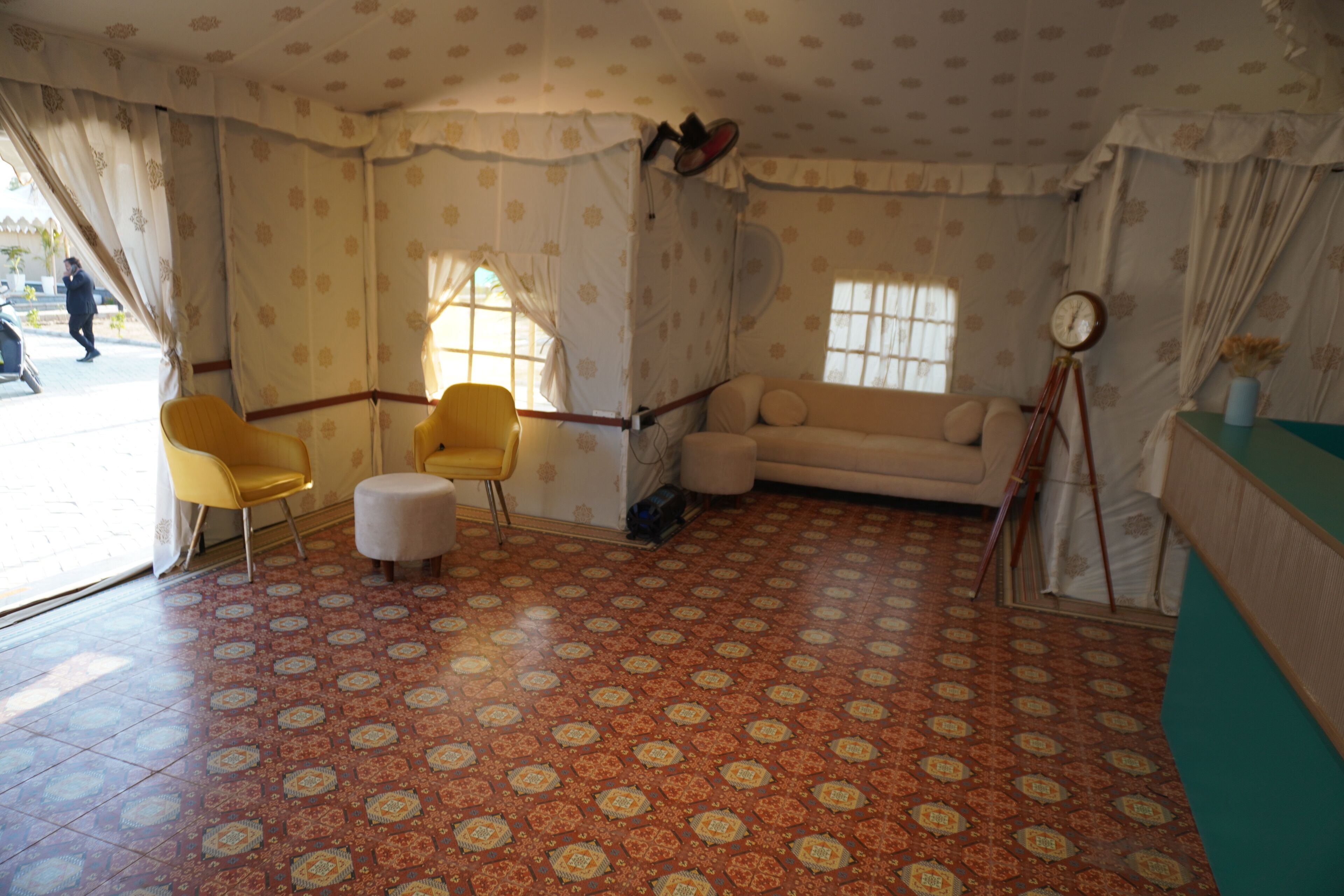 A room with patterned carpet, a sofa, two yellow chairs, a small ottoman, and a tripod with a clock.