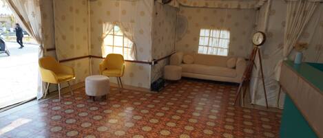 A room with patterned carpet, a sofa, two yellow chairs, a small ottoman, and a tripod with a clock.