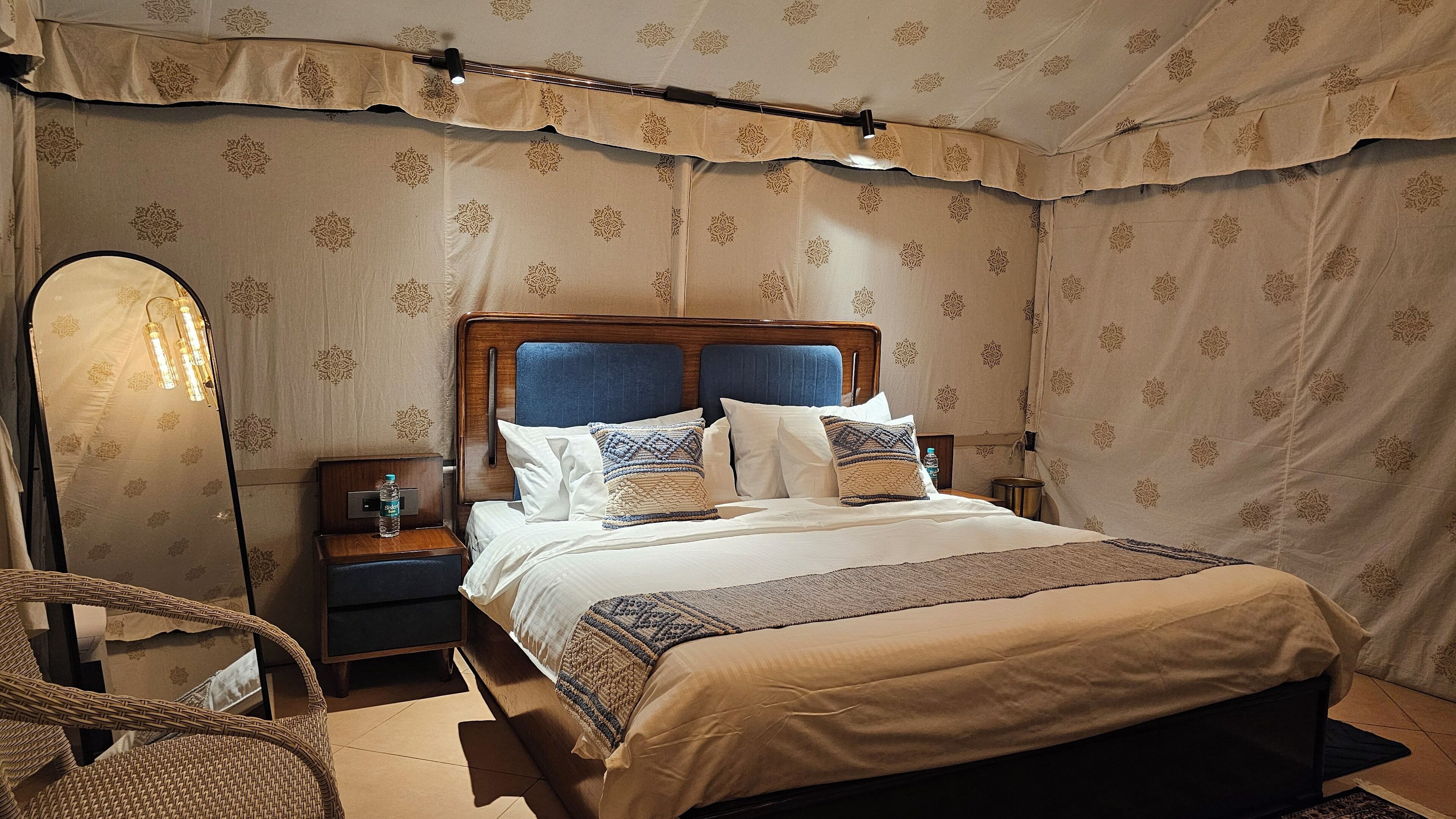 A bedroom with a wooden headboard, a bed with white and patterned bedding, a nightstand with a bottle, and a wicker chair.