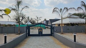 A gated entrance with a decorative gate, a paved pathway, and a white building with a peaked roof in the background. - Mayura Retreat (Danta Ramgarh)