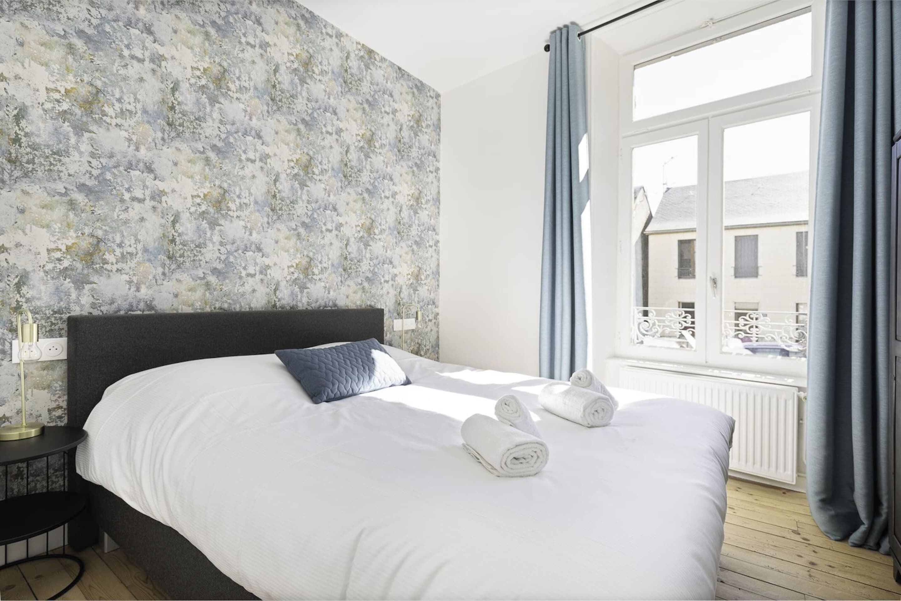A hotel room with a bed, a nightstand, a window with curtains, and a view of buildings outside.