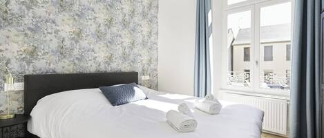A hotel room with a bed, a nightstand, a window with curtains, and a view of buildings outside.