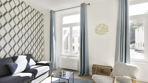 Ein modernes Wohnzimmer mit einem grauen Sofa, einem kleinen Tisch und einem Sessel. Ein Fenster mit Vorhängen, ein Heizkörper und eine Wand mit geometrischem Muster sind zu sehen.