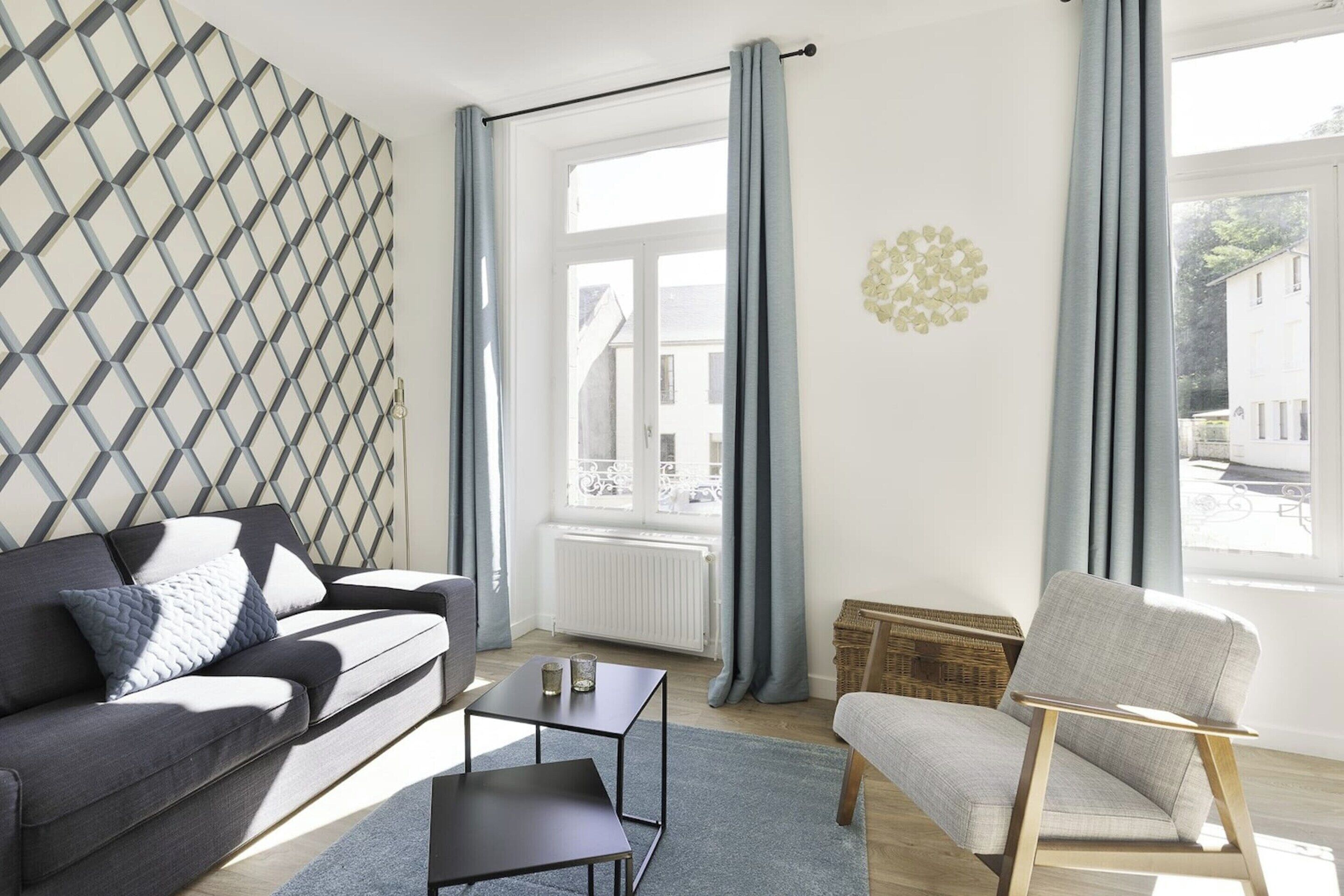 Ein modernes Wohnzimmer mit einer grauen Couch, einem kleinen schwarzen Tisch, einem hellgrauen Sessel und einer Wand mit Muster.
