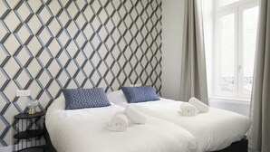 Ein Hotelzimmer mit Wand im geometrischen Muster, einem Bett mit weißer Bettwäsche, zwei Kissen, einem Nachttisch mit Lampe und einem Fenster mit Vorhängen.
