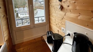 A small kitchen with wooden walls, a window with a view of a road and buildings, a toaster, a kettle, and a spoon hanging on the wall. - Comfortable Ski Stay in Mont-dore (Mont-Dore)